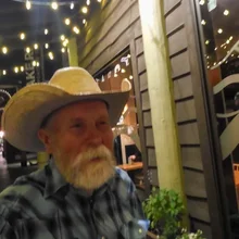 Older man with a white beard wearing a plaid shirt and cowboy hat standing outside a building decorated with string lights